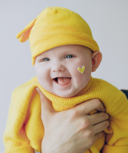 un bébé est en train de sourire, il porte un bonnet et un pull jaune canari et il a un coeur jaune sur la joue