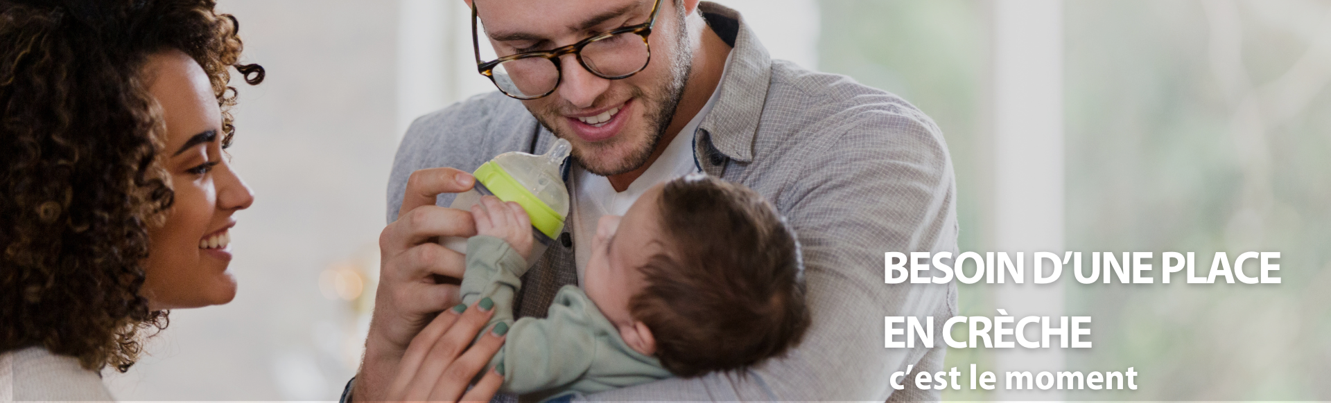 Un père tient son enfant dans les bras en souriant et va lui donner le biberon, la mère les regarde en souriant.